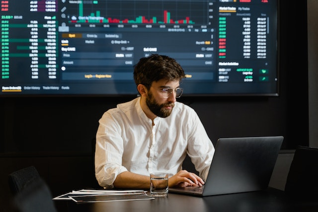 Analyst working on a laptop in front of a large screen of market charts and figures