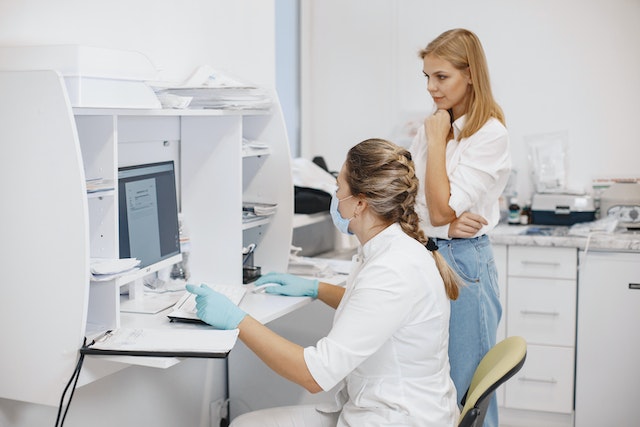 Two women in a clinic looking at a computer screen, one wearing a mask and gloves