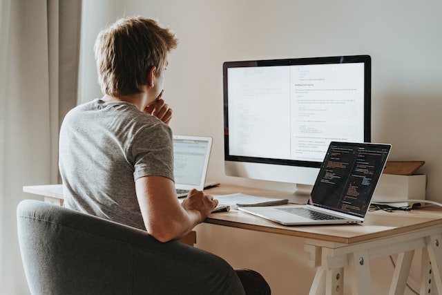 Developer at a desk studying documentation on a monitor, with code open on a laptop