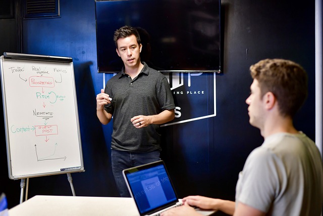 Man presenting a workflow diagram on a whiteboard to a colleague working on a laptop