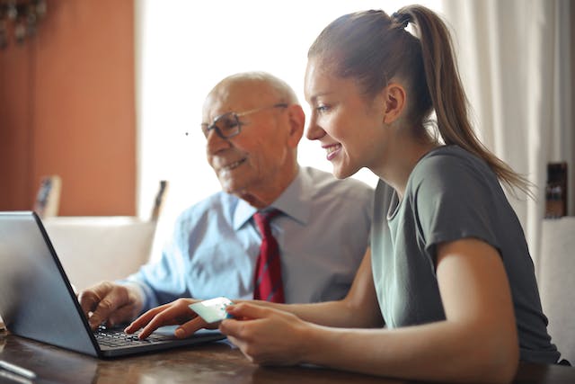 Young woman helping an older man use a laptop at a table while holding a bank card