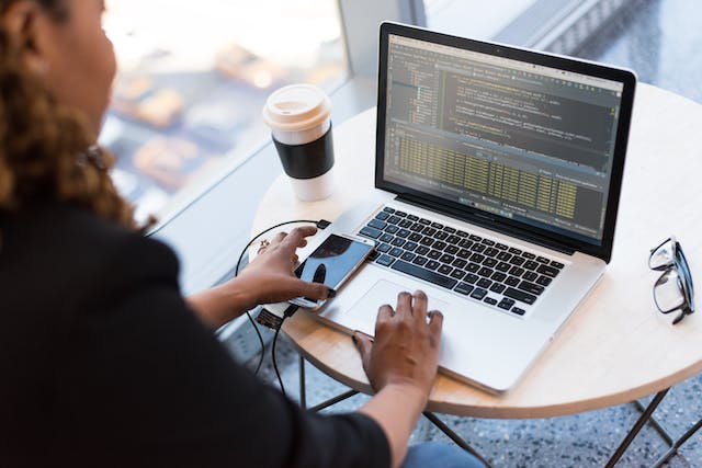 Developer at a café table working on a laptop with code while holding a phone
