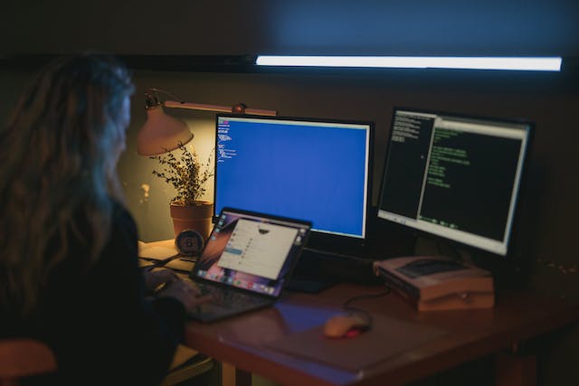 Woman working at a desk with two monitors and a laptop in a dimly lit room