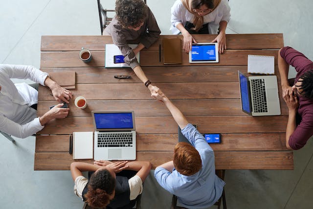 Overhead view of a team around a wooden table with laptops as two colleagues shake hands