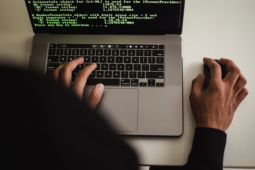 Hands at a laptop showing green console text, one hand typing and one on a mouse