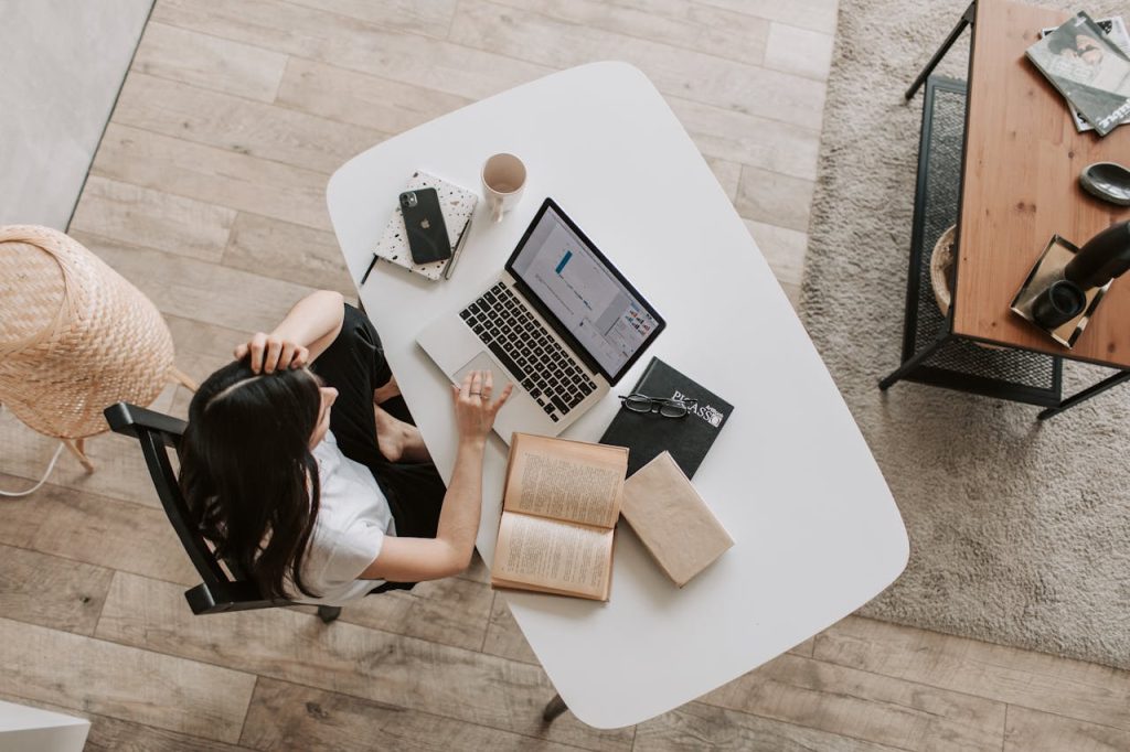 Overhead view of a woman working on a laptop at a white desk with books and a phone