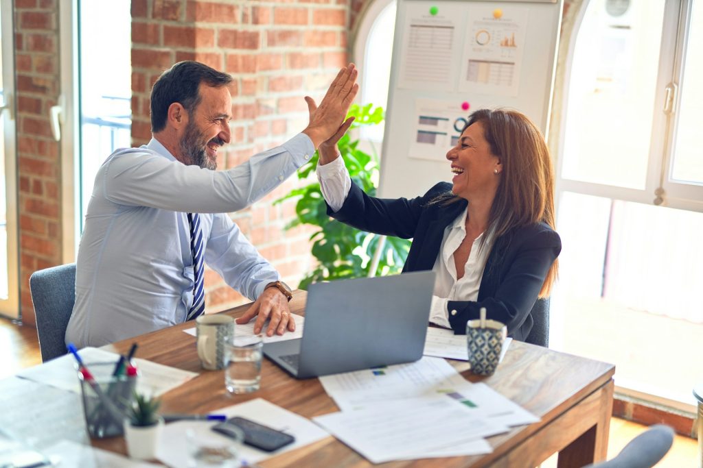 Two colleagues high-fiving across a desk with a laptop and paperwork