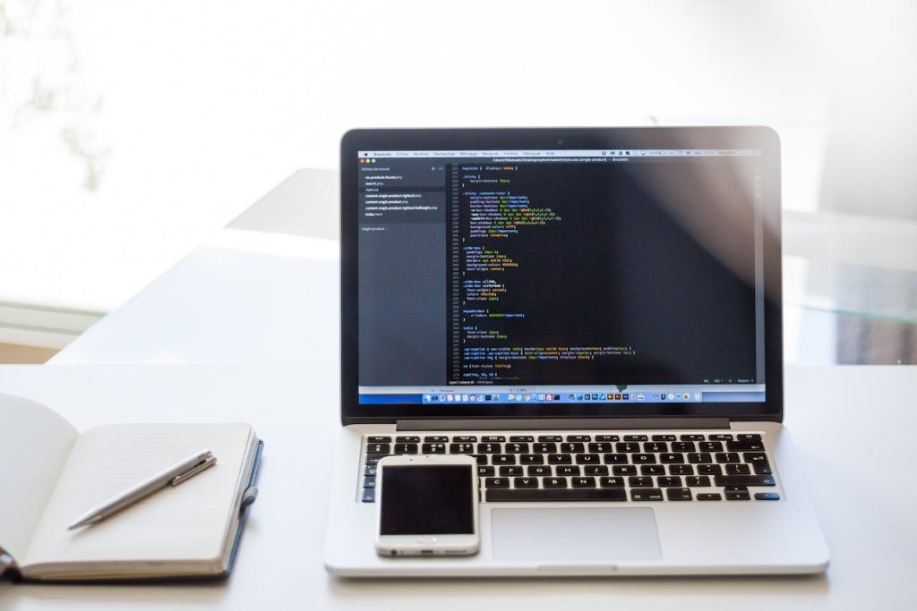 Laptop showing code on a white desk with a smartphone and an open notebook