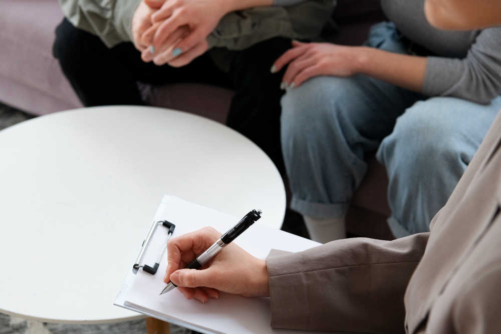 Therapist taking notes on a clipboard during a session with two people on a sofa